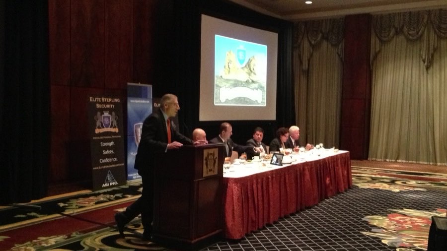 AJ Zabadne speaking at a lectern beside a head table at an Elite Sterling Security event